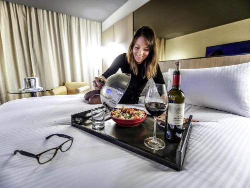 a woman sitting on a bed with a tray of food and wine at Pullman Bordeaux Lac in Bordeaux