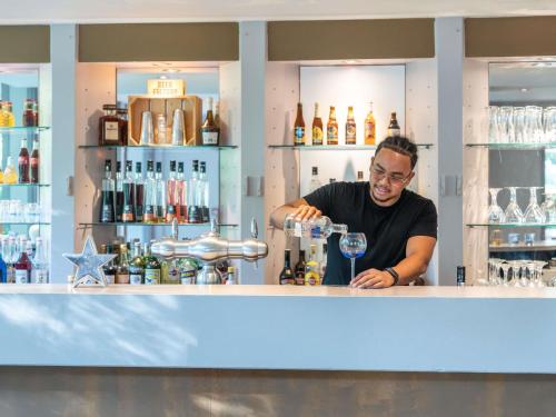 a man standing behind a bar with a glass at Mercure Besancon Parc Micaud - Hotel & Bar in Besançon