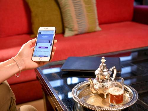a person holding a cell phone next to a coffee table with a tea kettle at Mercure Shéhérazade Rabat in Rabat
