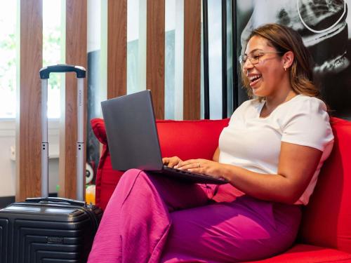 a woman sitting on a red couch with a laptop at ibis Joinville in Joinville