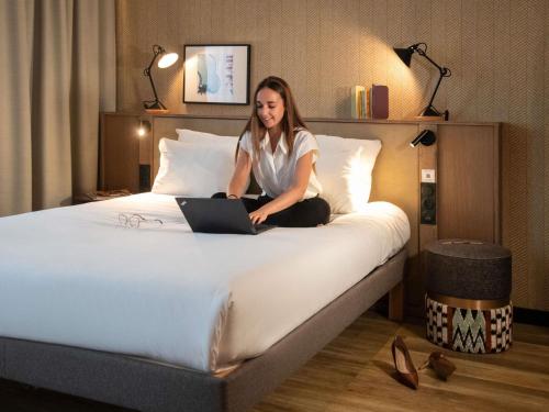 a woman sitting on a bed with a laptop at Novotel Suites Nice Airport in Nice