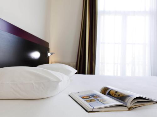 a book on a bed with a pillow and a window at Mercure Lille Roubaix Grand Hôtel in Roubaix