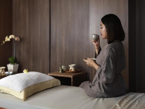 a woman sitting on a bed holding a cup of coffee at Novotel Bali Ngurah Rai Airport in Kuta