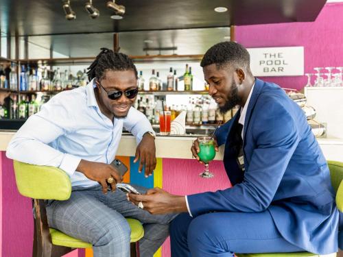 two men sitting at a bar looking at a cell phone at Ibis Styles Accra Airport in Accra