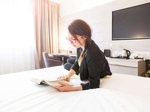 a woman sitting on a bed reading a book at Novotel Brescia Due in Brescia