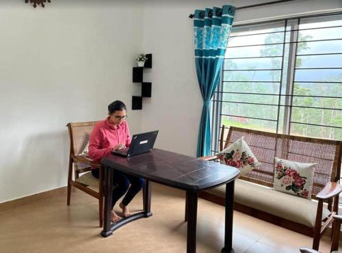 a woman sitting at a table with a laptop at Green Echoes by Tripture - Holiday Villas in Coorg in Cherambane