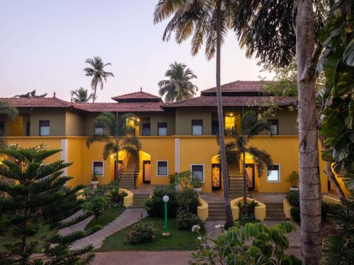 an exterior view of a yellow building with palm trees at Mercure Goa Devaaya Resort in Old Goa
