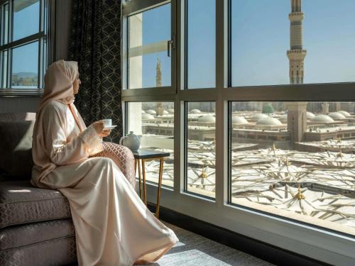 a woman sitting on a couch with a cup of coffee looking out a window at Anwar Al Madinah Mövenpick in Al Madinah