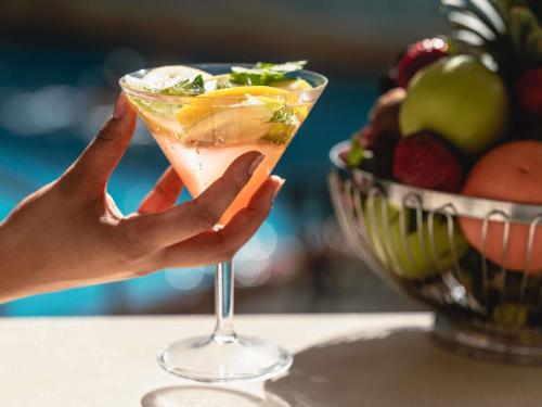 a person holding up a cocktail in front of a bowl of fruit at Adagio Jeddah City Center in Jeddah