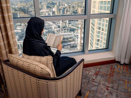 a woman sitting in a chair reading a book in a window at Swissotel Al Maqam Makkah in Makkah
