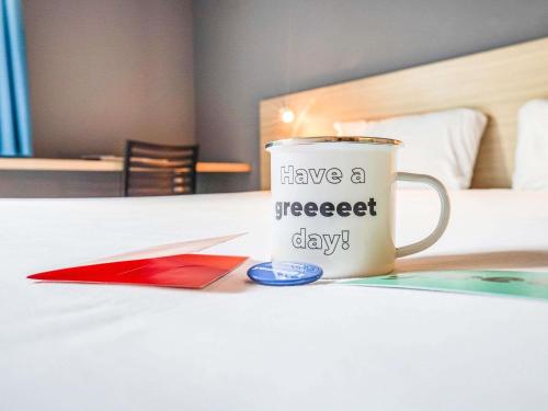 a coffee cup with have a sweet day written on it on a bed at greet Hotel Bordeaux Aeroport in Mérignac