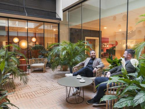 two men sitting in chairs in a lobby with plants at Ibis Styles Sevilla City Santa Justa in Seville