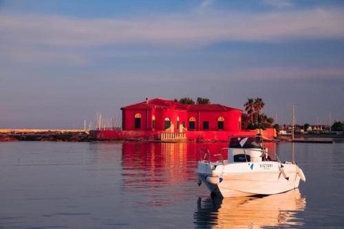 a boat in the water in front of a red house at Le Saline Rooms Marzamemi in Marzamemi