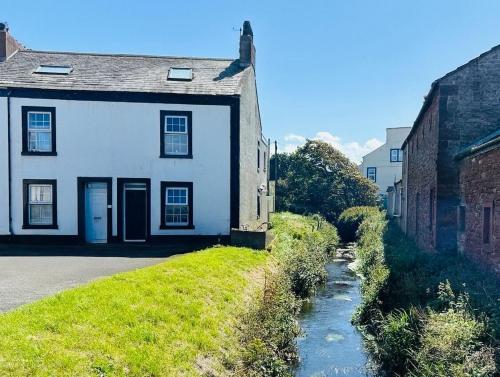 an old white house with a river in front of it at Old Post Office By The Sea in Allonby