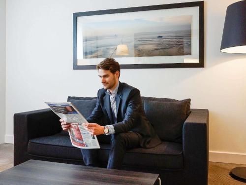 a man sitting on a couch reading a newspaper at Novotel Saclay in Saclay