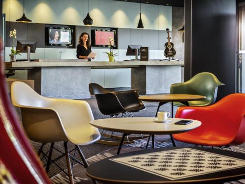 a bar with colorful chairs and a woman standing at a counter at Ibis Marne la Vallée Noisy in Noisy-le-Grand
