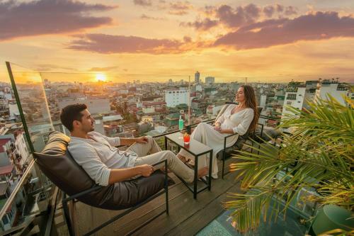 a man and woman sitting in chairs on a balcony with the sunset at Kecho Legacy Hotel Hanoi & Spa in Hanoi