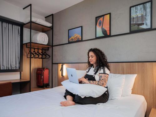 a woman sitting on a bed with a laptop at Ibis Foz do Iguaçu in Foz do Iguaçu