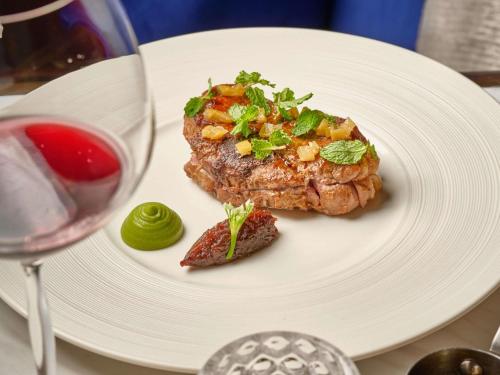 a white plate with a piece of meat and a glass of wine at Hotel Des Arts Saigon Mgallery Collection in Ho Chi Minh City