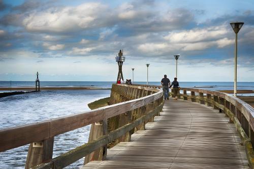 two people walking down a wooden boardwalk towards the ocean at Courseulles Comme À La Maison in Courseulles-sur-Mer