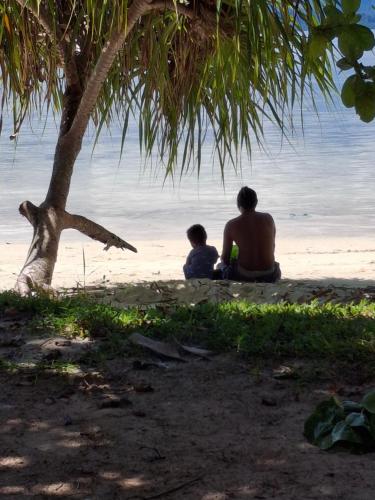 a man and a child sitting under a tree on the beach at Eddo Homestay in Kri