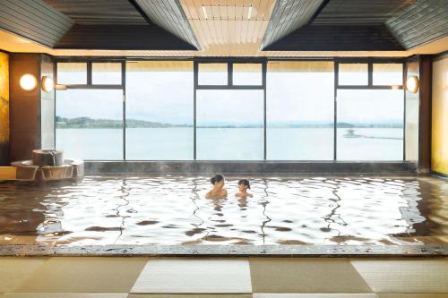 two people in the water in a pool with windows at Ooedo Onsen Monogatari Premium Kaga Maruya in Kaga