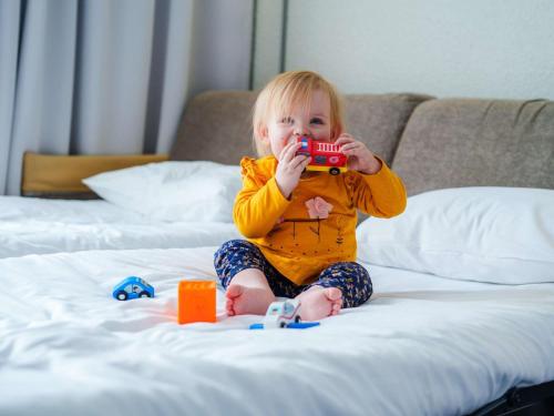 a little girl sitting on a bed holding a red camera at Novotel Saint Avold in Saint-Avold