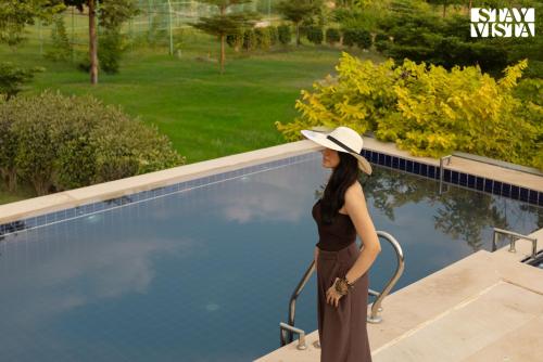 Una mujer con sombrero parada junto a una piscina. en StayVista at The Stone Age Home with Pool, Lawn, BBQ & Bonfire, en Jaipur