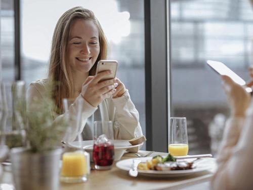 Uma mulher sentada numa mesa a olhar para o telemóvel. em Mercure Hotel Koeln Belfortstrasse em Colónia