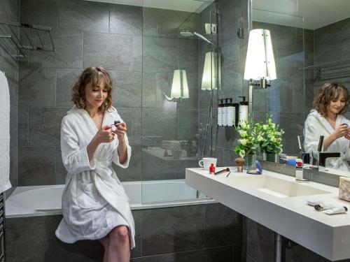 a woman sitting in a bathroom looking at her cell phone at Sofitel Strasbourg Grande Ile in Strasbourg