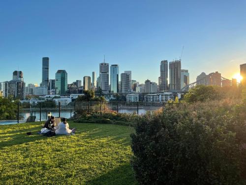 two people sitting on the grass in a park with a city skyline at Mid-Century Luxe - Story Bridge & River Views in Brisbane