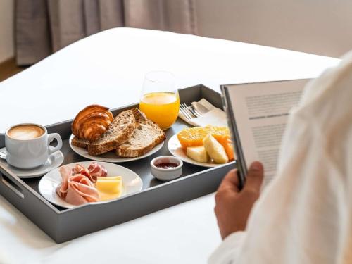 Una bandeja de alimentos para el desayuno con una persona leyendo un libro. en Costanero Hotel Montevideo - MGallery Collection, en Montevideo