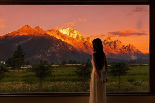 a woman standing in front of a window looking at the mountains at ShanJuanYun Panoramic Sunlit Yulong Snow Mountain Hotel in Baisha