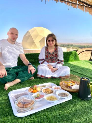 a man and woman sitting on the grass with a tray of food at Aman dome desert in Ḩawīyah