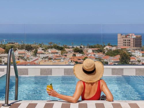 a woman sitting next to a swimming pool with a drink at Elkonin Tel Aviv - MGallery Hotel Collection in Tel Aviv