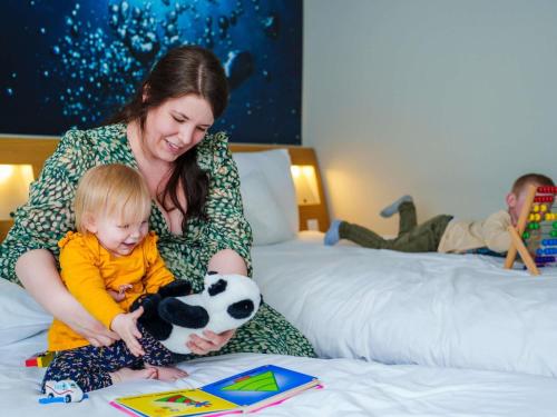 a woman sitting on a bed with a child holding a stuffed animal at Novotel Saint Avold in Saint-Avold