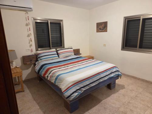 a bedroom with a bed with a striped comforter and two windows at הבית במושב in Umm el Shuf