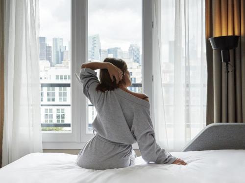 a woman sitting on a bed looking out a window at Aparthotel Adagio Paris Suresnes Longchamp in Suresnes
