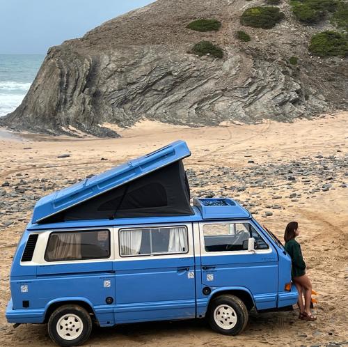 Una mujer parada junto a una camioneta azul y blanca en una playa. en rent a campervan vw t3 azul in Algarve Portugal, en Bicas