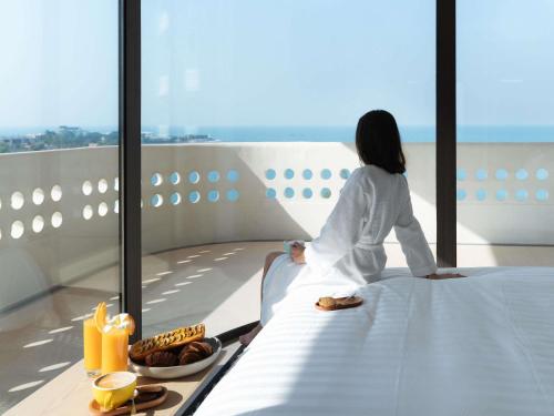 una mujer sentada en una cama mirando por la ventana en Novotel Sihanoukville Holiday Resort, en Sihanoukville