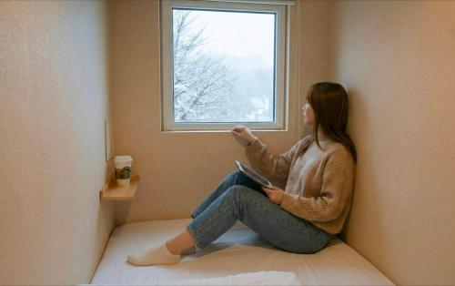 a woman sitting on a bed looking out of a window at Sho inn MINIMAL HOTEL 小樽駅から無料送迎あり in Otaru