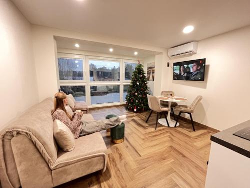 a woman sitting on a couch in a living room with a christmas tree at Tėčio ir Dukters pajūrio apartamentai - Šventojoje in Palanga