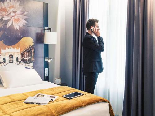 a man in a suit talking on a cell phone next to a bed at Mercure Nancy Centre Place Stanislas in Nancy