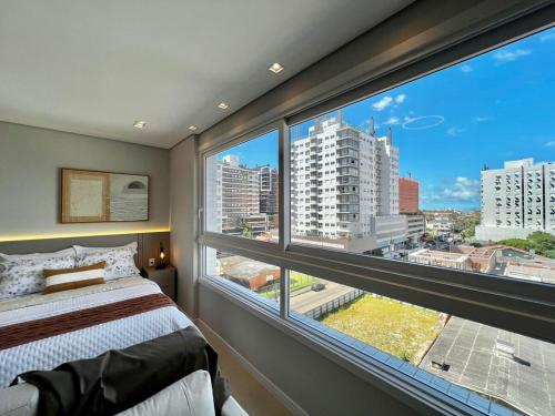 a bedroom with a large window with a view of a city at Studio Capão - novo com piscina e rooftop in Capão da Canoa