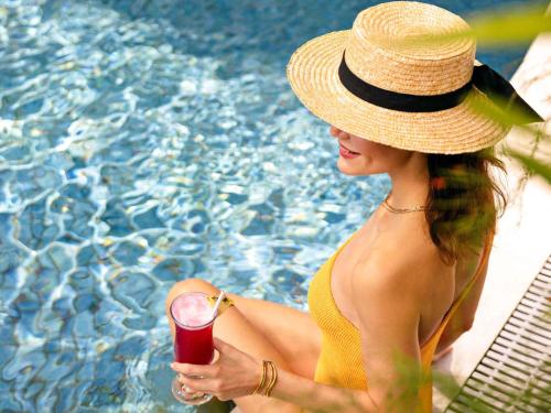 a woman in a hat holding a drink next to a pool at Fairmont Singapore in Singapore