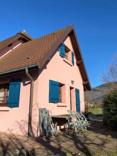 Gîte Dreiviertelacker في Husseren-Wesserling: منزل وردي اللون ذو مصاريع زرقاء وطاولة وكراسي
