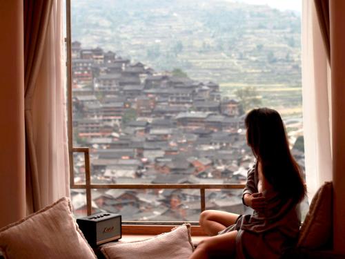 a woman sitting on a window sill looking at a city at Seven Letters Preface Hotel Xijiang Qianhu Miao Village in Xijiang