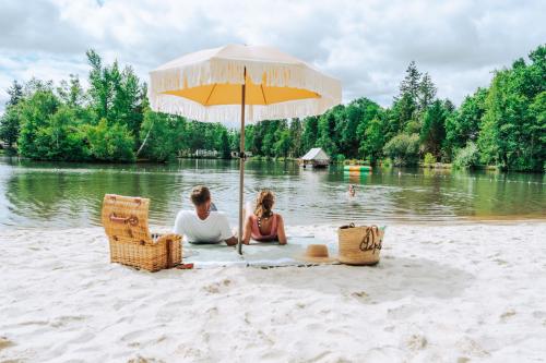 two people sitting on a beach under an umbrella at Camping Etang de Sologne in Nouan-le-Fuzelier