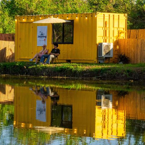 two people sitting under an umbrella next to a body of water at Lakeside Cargo in Yellow - A Cozy Container Stay in San Pa Tong