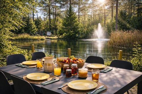 a table with plates of food and a bowl of fruit next to a lake at StayatSas Pippa grote omheinde tuin in het bos aan grote vijver, zwembad en restaurant op het park, stijlvol ingericht,veel privacy in Doornspijk